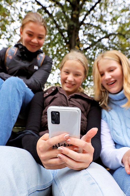 Tre ungdomar sitter ute med ett träd i bakgrunden och skrattar och tittar på en mobiltelefon.
